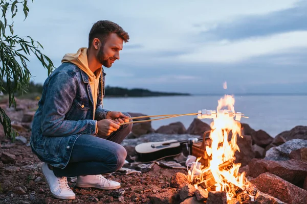 Hombre feliz tostando malvaviscos en palos cerca de la hoguera - foto de stock