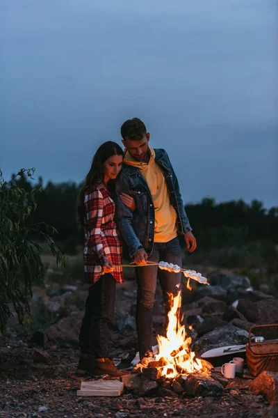 Couple standing near burning bonfire and roasting marshmallows on sticks — Stock Photo