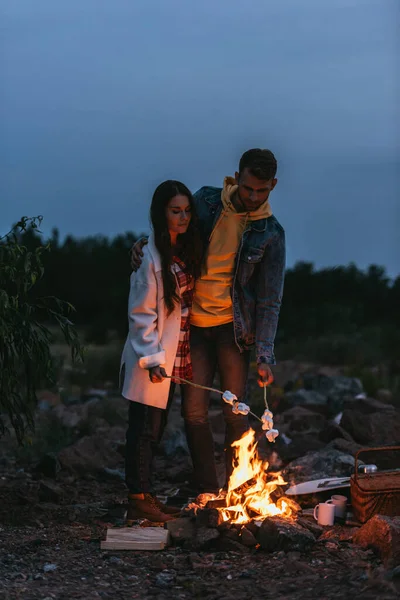 Couple standing near bonfire and roasting marshmallows on sticks — Stock Photo