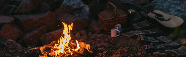 Panoramic crop of burning bonfire near plaid blanket, wicker basket, cups and acoustic guitar — Stock Photo