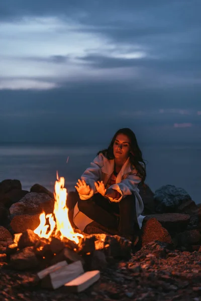Enfoque selectivo de la mujer calentándose cerca de la hoguera en llamas por la noche - foto de stock