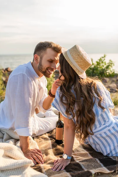 Hombre feliz tocando la cara de la mujer alegre en sombrero de paja cerca de botella con vino - foto de stock