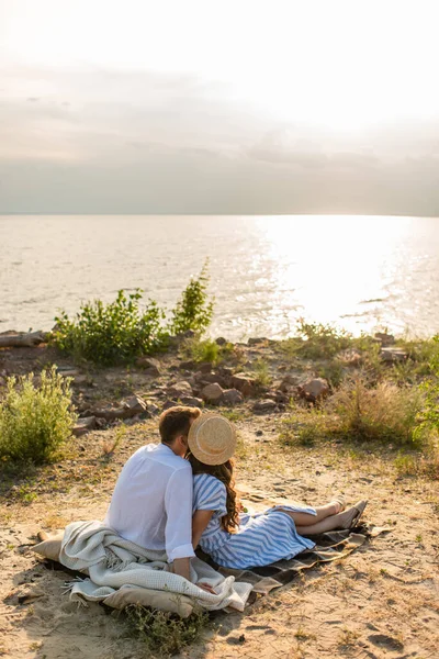 Vista trasera de la joven mujer en sombrero de paja y el hombre sentado en la manta cerca del río - foto de stock