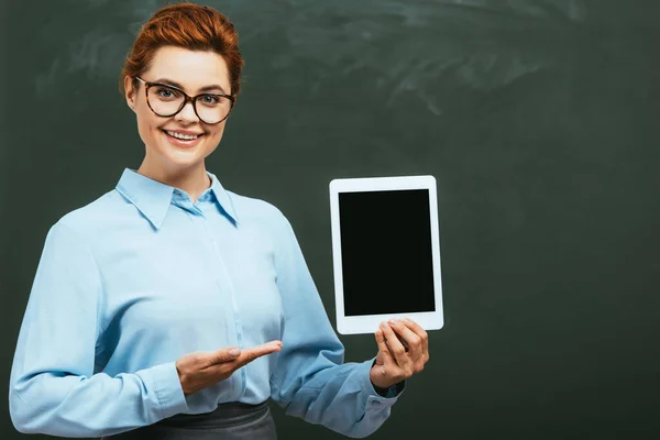 Enseignant heureux pointant avec la main à la tablette numérique avec écran blanc près du tableau noir — Photo de stock