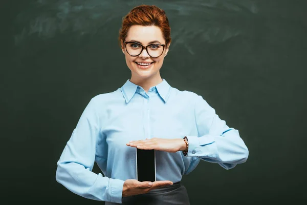 Attractive, happy teacher holding smartphone with blank screen while standing near chalkboard — Stock Photo