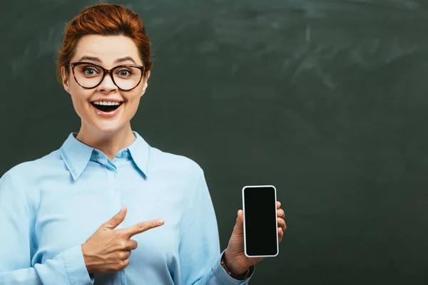 Happy, beautiful teacher pointing at smartphone with blank screen while standing near chalkboard — Stock Photo