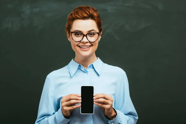 Happy, attractive teacher showing smartphone with blank screen while standing near chalkboard — Stock Photo