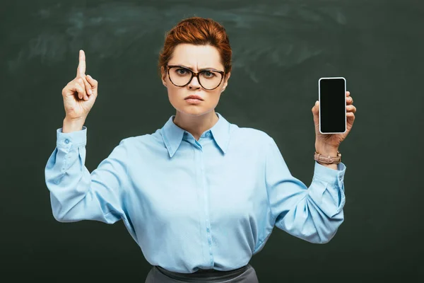 Strict teacher showing attention gesture while showing smartphone with blank screen near chalkboard — Stock Photo
