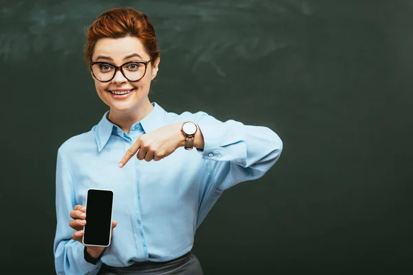 Profesor feliz y atractivo apuntando con el dedo al teléfono inteligente con pantalla en blanco cerca de pizarra - foto de stock