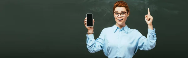 Horizontal concept of smiling teacher showing idea gesture while holding smartphone with blank screen near chalkboard — Stock Photo