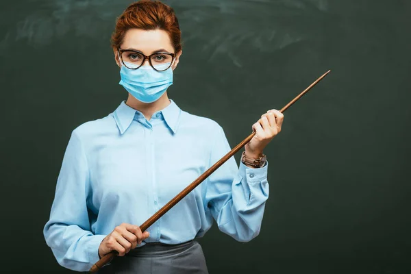 Young teacher in medical mask looking at camera while holding pointing stick near blank chalkboard — Stock Photo