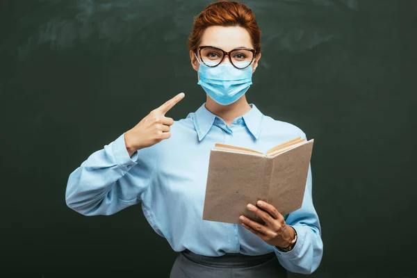 Giovane insegnante con libro aperto che punta alla maschera medica mentre in piedi vicino alla lavagna — Foto stock