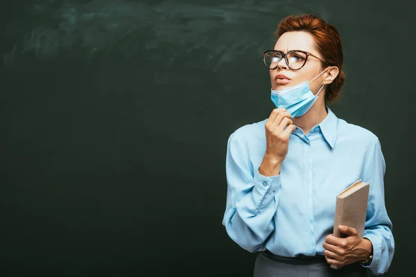 Insegnante con libro toccare maschera medica mentre in piedi vicino lavagna e guardando altrove — Foto stock