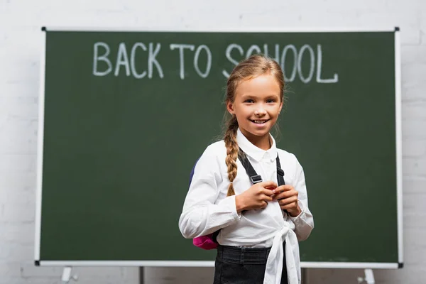 Enfoque selectivo de colegiala adorable con mochila sonriendo a la cámara cerca de pizarra con inscripción de vuelta a la escuela - foto de stock