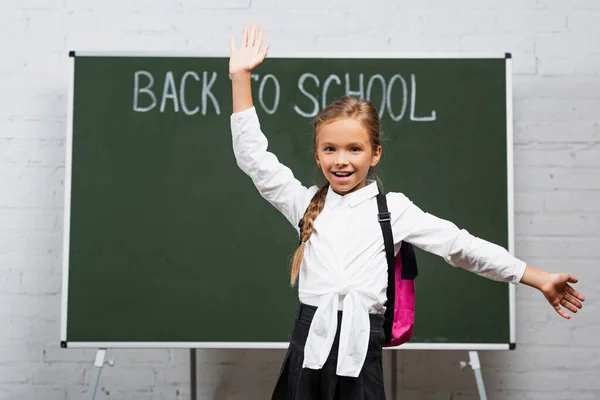 Селективный фокус счастливой школьницы с рюкзаком и открытыми объятьями, улыбающейся в камеру возле доски с надписью 