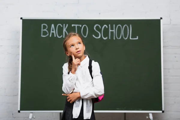 Écolière réfléchie touchant le visage et regardant vers le haut près de tableau noir avec inscription de retour à l'école — Photo de stock