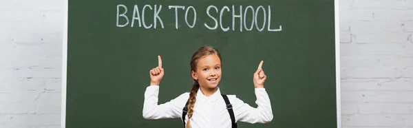 Horizontales Konzept des glücklichen Schulmädchens, das mit dem Finger auf die Schulaufschrift auf der Tafel zeigt — Stockfoto