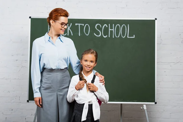 Happy teacher and schoolgirl standing near chalkboard with back to school lettering — Stock Photo