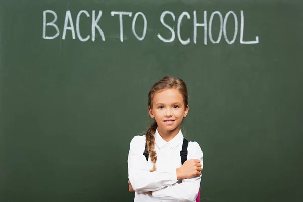 Scolaretta sorridente in piedi con le braccia incrociate vicino alla lavagna con iscrizione di ritorno a scuola — Foto stock