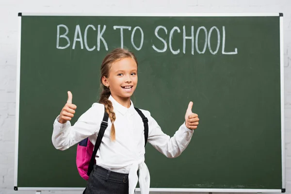 Colegiala feliz con mochila mostrando los pulgares hacia arriba cerca de pizarra con letras de vuelta a la escuela - foto de stock