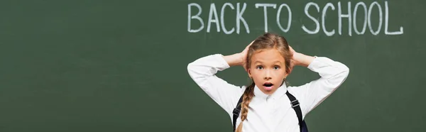 Plan panoramique d'écolière choquée touchant la tête près du tableau noir avec inscription de retour à l'école — Photo de stock