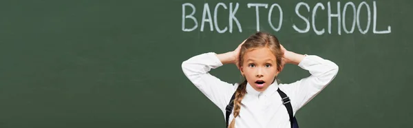 Panoramic shot of shocked schoolgirl touching head near chalkboard with back to school inscription — Stock Photo