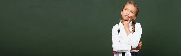 Horizontal concept of thoughtful schoolgirl touching face and looking up near chalkboard — Stock Photo