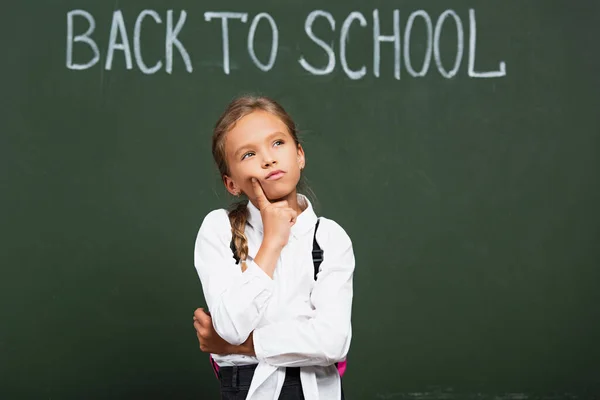 Écolière réfléchie toucher le visage et regarder vers le haut près de tableau noir avec retour à l'école lettrage — Photo de stock