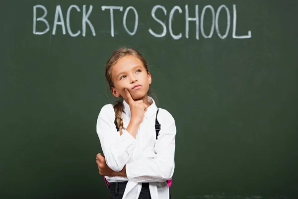 Reflexiva colegiala tocando la cara y mirando hacia arriba cerca de pizarra con letras de vuelta a la escuela - foto de stock