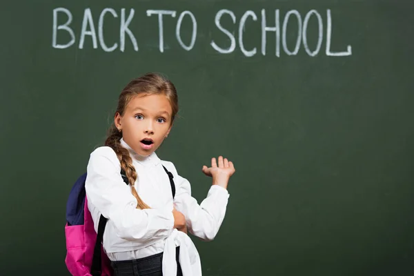 Colegiala sorprendida con mochila apuntando con la mano a la pizarra con inscripción de vuelta a la escuela - foto de stock