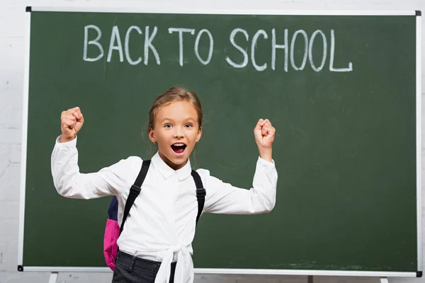 Aufgeregtes Schulmädchen zeigt Siegergeste neben Kreidetafel mit Schulschriftzug — Stockfoto