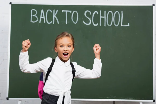 Studentessa eccitato mostrando gesto vincitore vicino lavagna con torna a scuola lettering — Foto stock