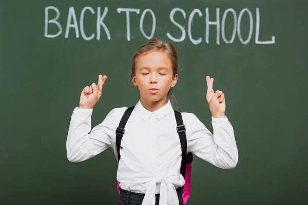 Adorable écolière avec les yeux fermés tenant les doigts croisés près de l'inscription de retour à l'école sur tableau — Photo de stock