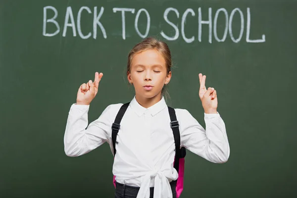 Adorabile studentessa con gli occhi chiusi tenendo le dita incrociate vicino alla iscrizione della scuola sulla lavagna — Foto stock