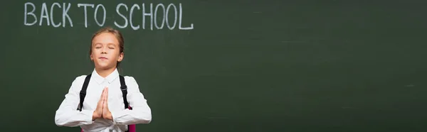 Concept panoramique d'écolière mignonne debout avec les mains priantes près du tableau noir avec lettrage de retour à l'école — Photo de stock