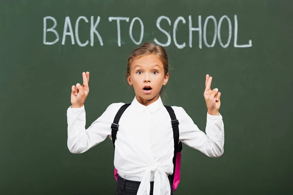 Shocked schoolgirl holding crossed fingers near back to school inscription on chalkboard — Stock Photo