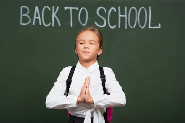 Mignonne écolière avec les yeux fermés tenant les mains priantes près de retour à l'école lettrage sur tableau noir — Photo de stock