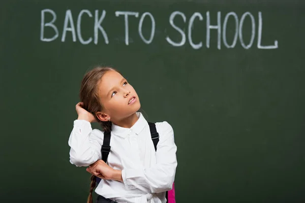 Messa a fuoco selettiva della studentessa pensosa che tocca la testa mentre guarda verso l'alto vicino alla lavagna con iscrizione di nuovo a scuola — Foto stock