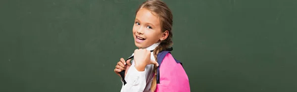 Horizontal concept of happy schoolgirl pointing with thumb at backpack near chalkboard — Stock Photo