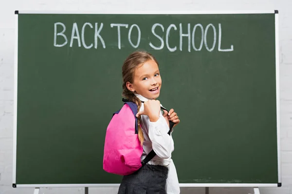 Cheerful schoolgirl pointing with thumb at backpack near chalkboard with back to school lettering — Stock Photo