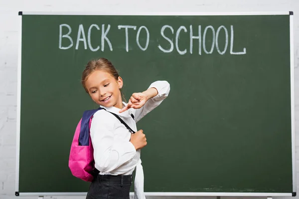 Colegiala sonriente apuntando con el dedo a la mochila cerca de pizarra con inscripción de vuelta a la escuela - foto de stock