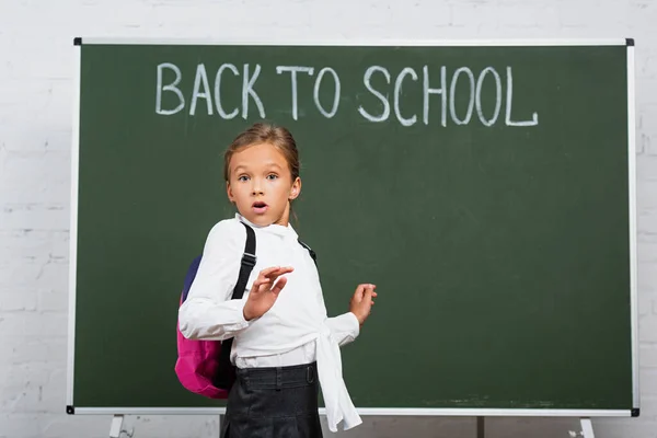 Shocked schoolgirl with backpack looking at camera near chalkboard with back to school inscription — Stock Photo