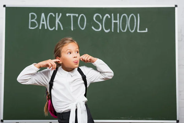 Astuta studentessa tappando le orecchie con le dita e sporgendo la lingua vicino alla lavagna con le lettere per tornare a scuola — Foto stock