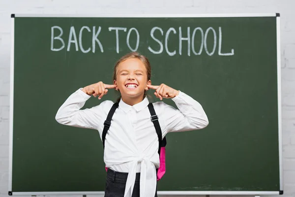 Displeased schoolgirl plugging ears with fingers near chalkboard with back to school inscription — Stock Photo