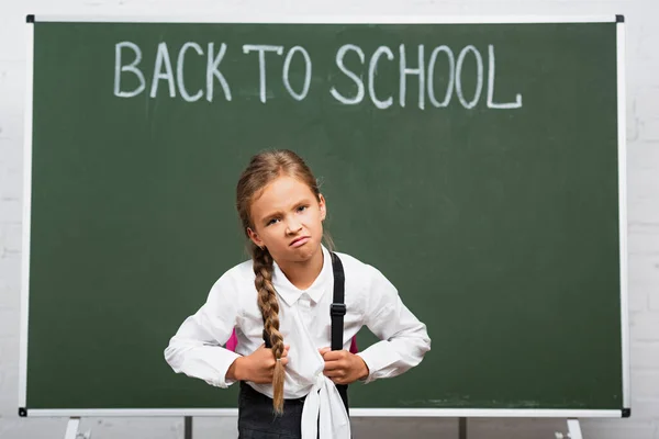 Displeased schoolgirl with heavy backpack near chalkboard with back to school lettering — Stock Photo