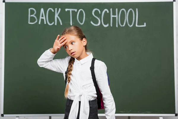 Colegiala agotada tocando la frente cerca de pizarra con letras de vuelta a la escuela - foto de stock