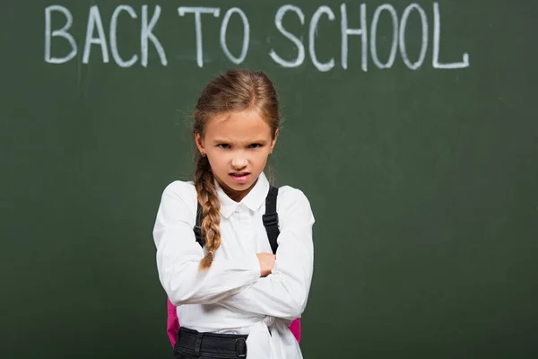 Angry schoolgirl standing with crossed arms near chalkboard with back to school lettering — Stock Photo