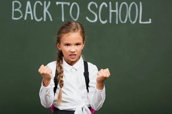 Selective focus of angry schoolgirl looking at camera and showing clenched fists near chalkboard with back to school lettering — Stock Photo