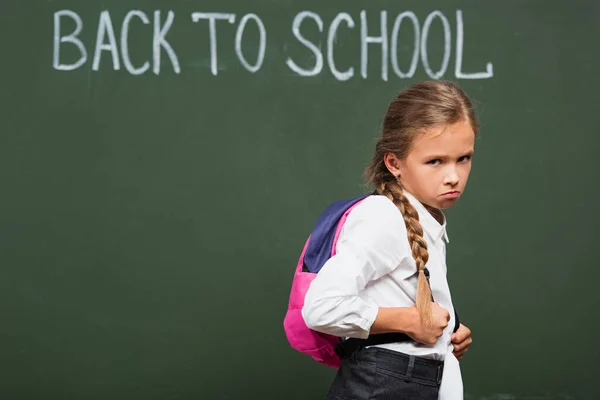 Upset schoolgirl looking at camera near chalkboard with back to school inscription — Stock Photo