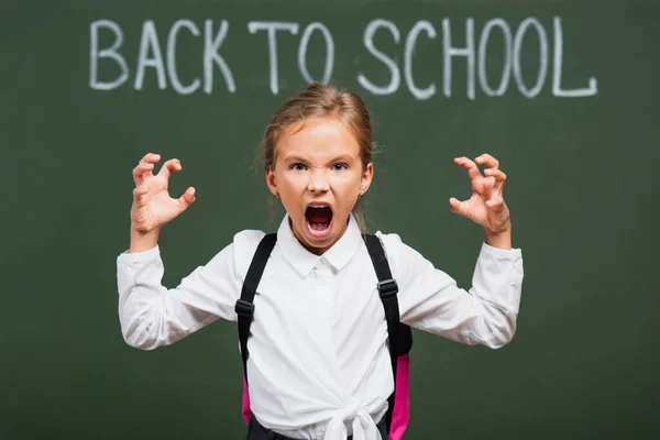 Selective focus of angry schoolgirl showing scaring gesture near chalkboard with back to school lettering — Stock Photo